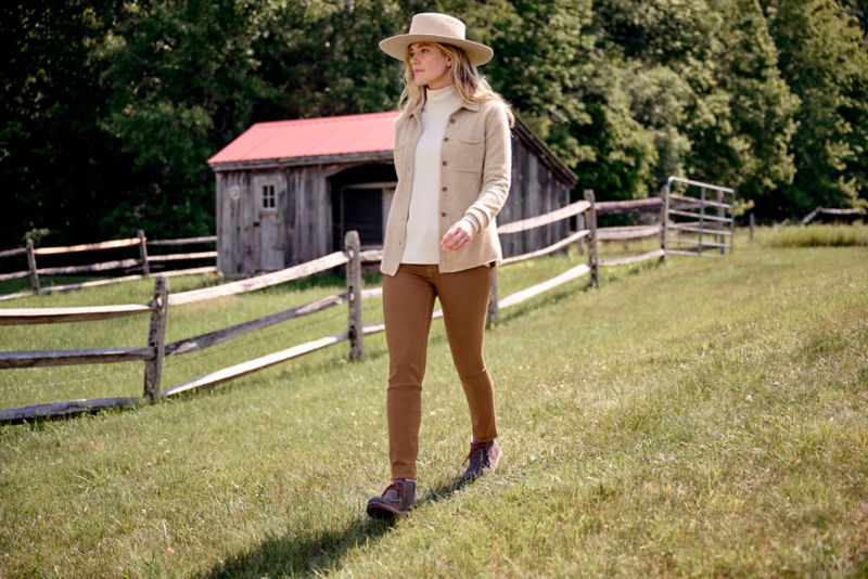 Woman in Boiled Cashmere Sweater Jacket walks along a wooden fencepost in a field.