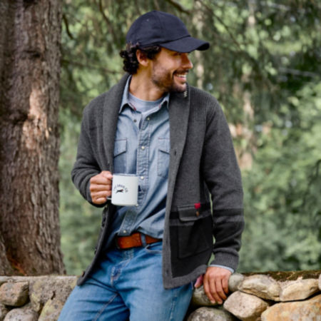 A man in the woods holding a mug with curly black hair wearing a gray cardigan.