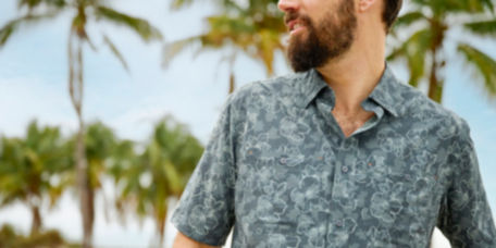 A man wearing a button-down printed teal shirt pauses mid-hike among tropical trees.