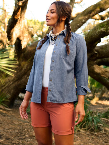 A woman wearing a blue chambray shirt and red shorts hiking through trees.