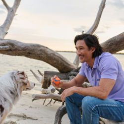 A man wearing a purple shirt at the beach throwing a ball to a white dog