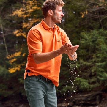 Man in Tangerine Orvis Signature Polo cleans his hands in the water.