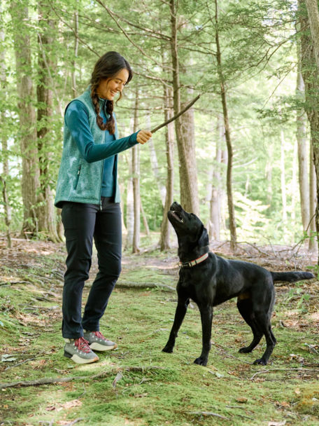 Woman in R65™ Sweater Fleece Vest plays with her dog while on a hike in the woods.