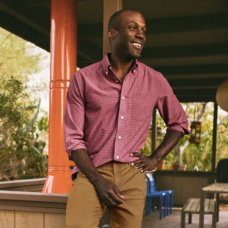 A man wearing a button down shirt and 5 pocket pant smiling and leaning on an outdoor patio.