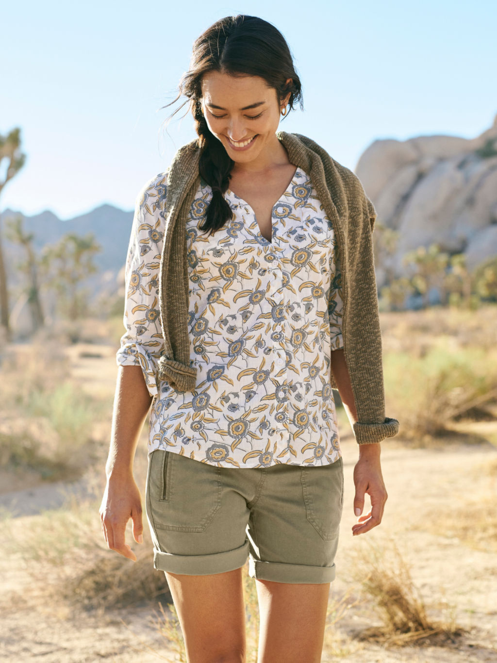 A model wearing a floral print shirt in white with yellow and grey flowers, olive khaki shorts, and a dark olive textured sweater draped over her shoulders