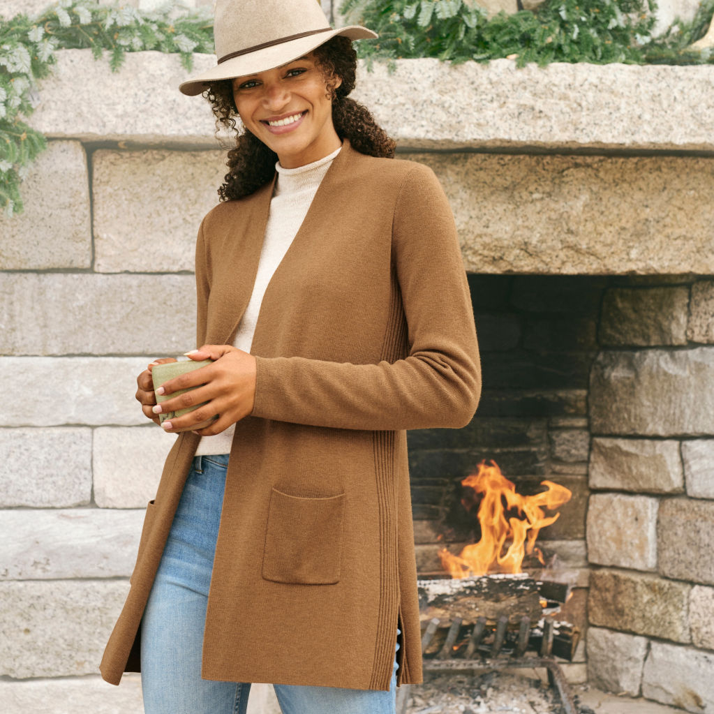 A model in a long tan cardigan poses in front of a huge stone fireplace 