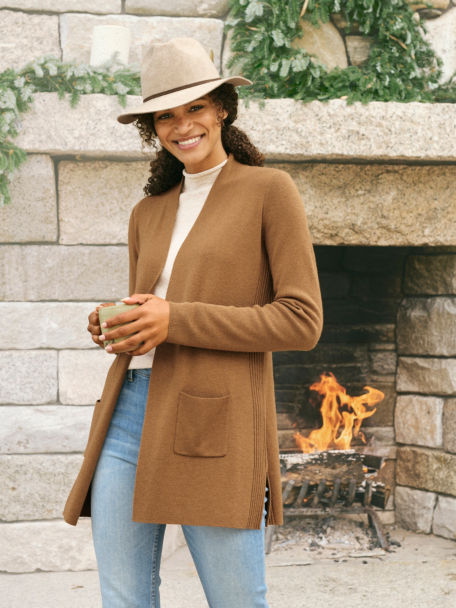 Woman in Dark Vicuna AnyWear Cardigan holds her coffee as she stands next to a lit rock fireplace.