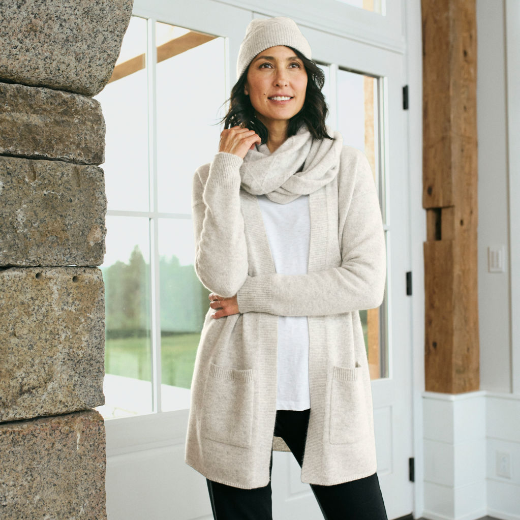 A model dressed in oatmeal-coloured cashmere cardigan, scarf, and hat stands next to a large window