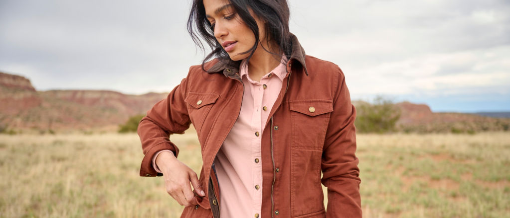 A hiker in a pink corduroy shirt and rust jacket opens a snap pocket while standing in a field.