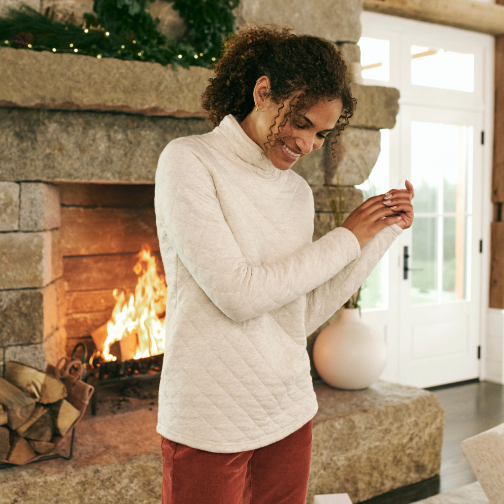 A model in an oatmeal quilted mockneck and rust corduroy pants warms herself in front of a stone fireplace
