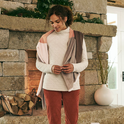 A model in an oatmeal-colored quilted top and multi-colored scarf walks in front of a stone fireplace.