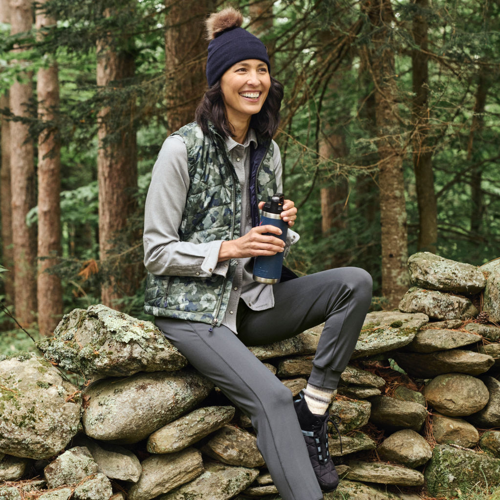 A model wearing a camo quilted vest sits on a stone wall in the woods