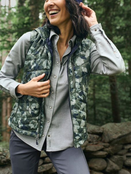 Woman in Sagebrush Mid Mountain Flannel Shirt and Juniper Painter Camo Recycled Drift Vest walks amongst pine trees.