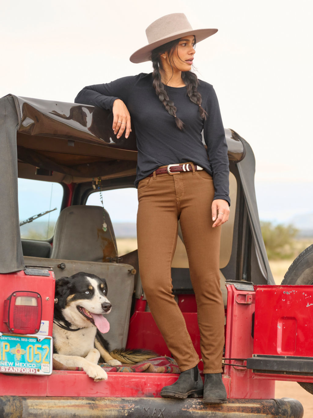 A woman stands on the rusty bumper of a red jeep while a black and white dog lays next to her.