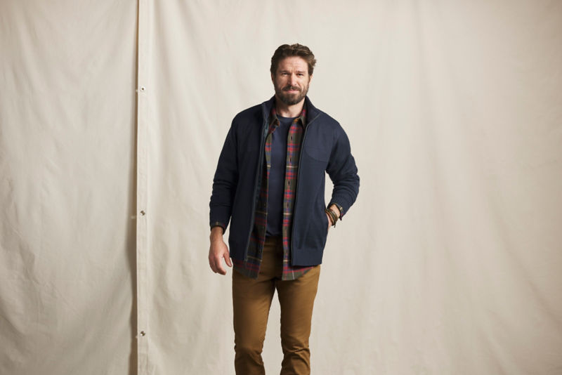 A model with his hand in the pocket of his navy blue jacket over a plaid shirt and brown pants.