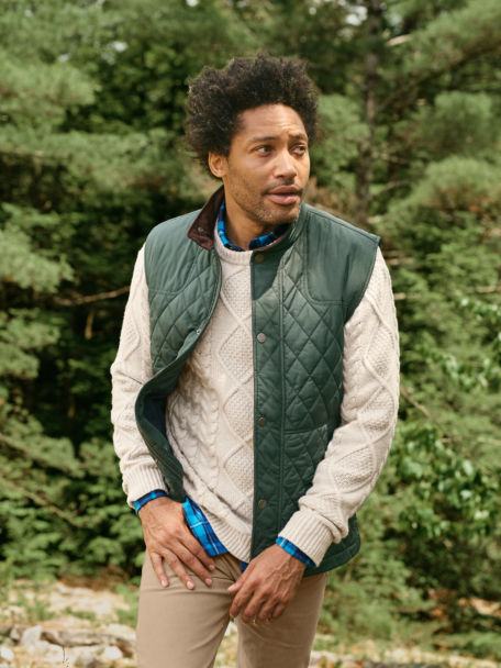 Man in Dark Pine RT7 Quilted Vest, Blue Fog Lodge Flannel Long-Sleeved Shirt, and Natural Cable Crewneck Sweater walks along a dusty trail in pine woods.