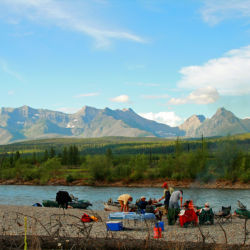 A group of people enjoy a river side lunch with mountains in the background.