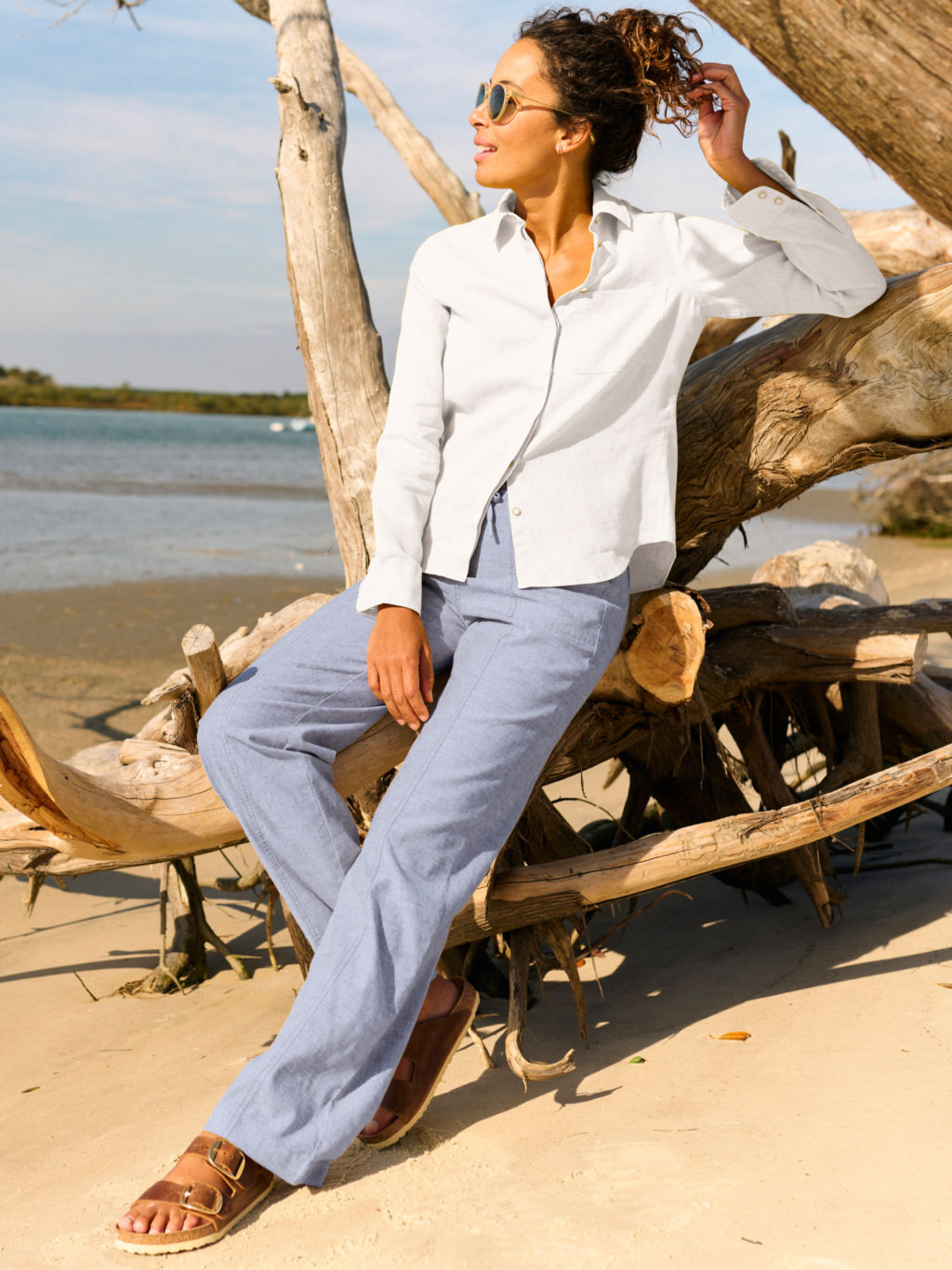 A woman sits on a pile of driftwood wearing blue linen pants with birkenstock sandals.