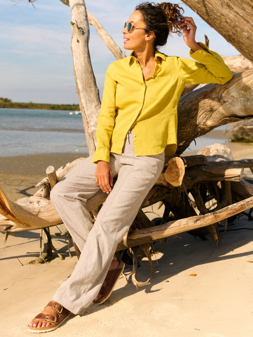 A model wearing a yellow button-down linen shirt and wide-legged linen pants lounges against driftwood on a beach.