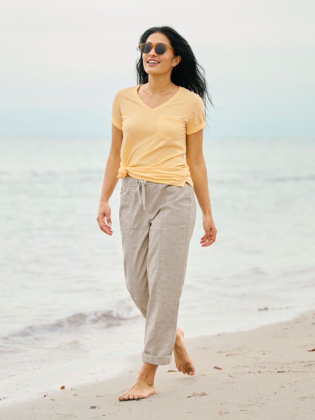 A woman walks along the beach wearing linen pants and a yellow t-shirt in a knot at the waist.