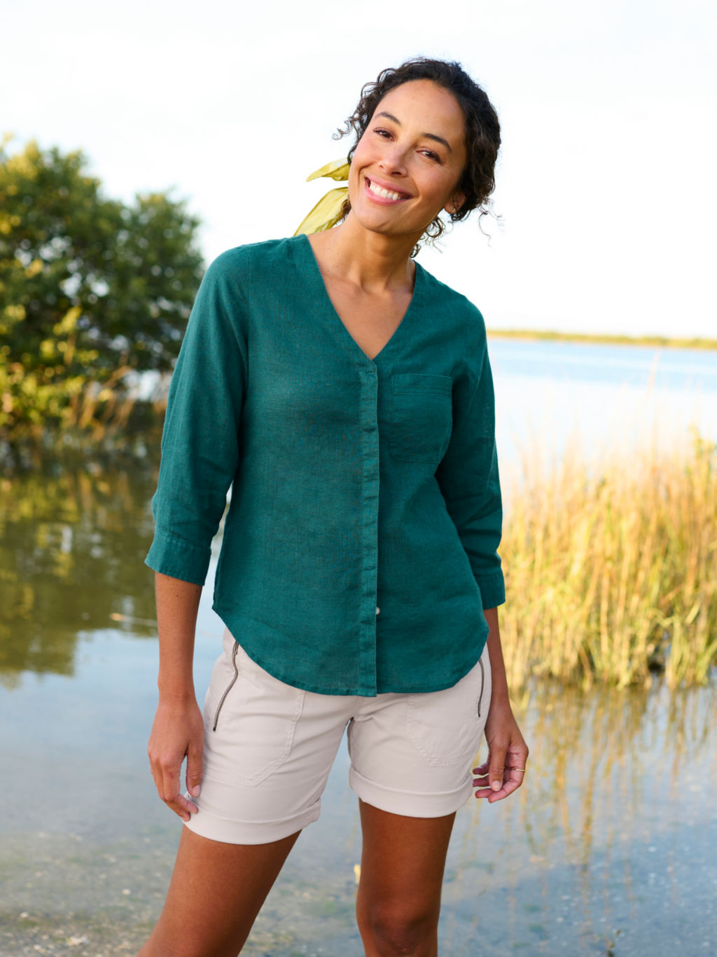A model wearing a dark green linen shirt and light khaki shorts stands at the edge of a marsh.
