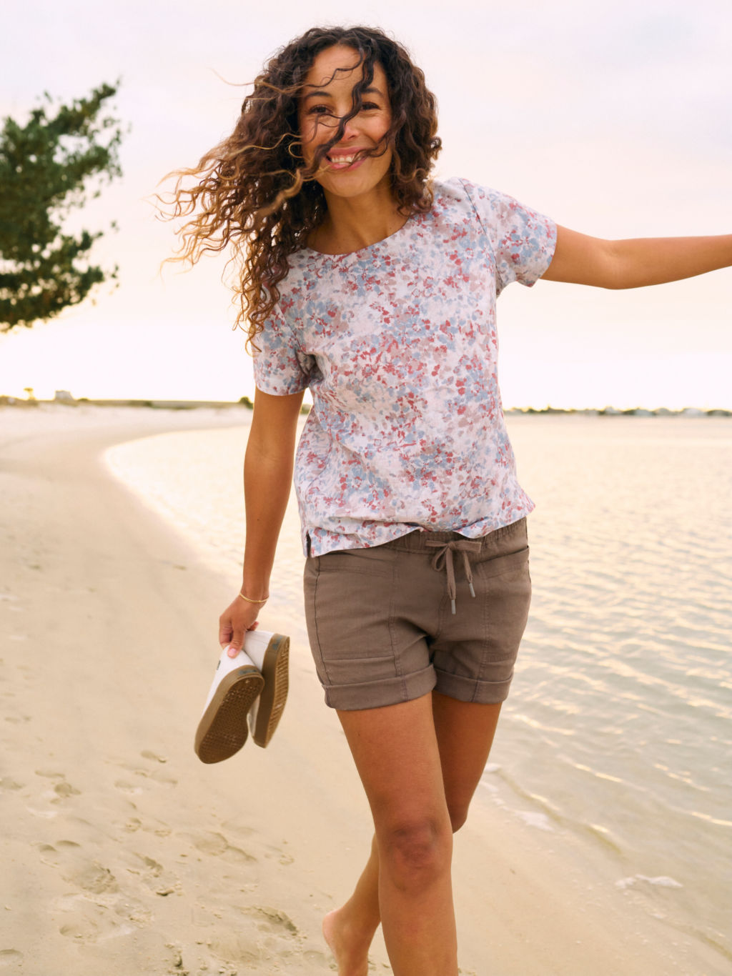 A model wearing a floral print linen t-shirt and brown colored shorts walks along the edge of the water on a sandy beach.