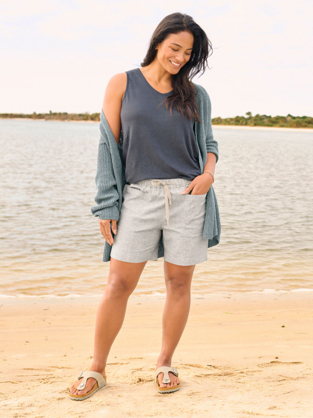 A model wearing a linen shorts, a navy sleeveless tee, and a seafoam green cardigan stands on a sandy beach.