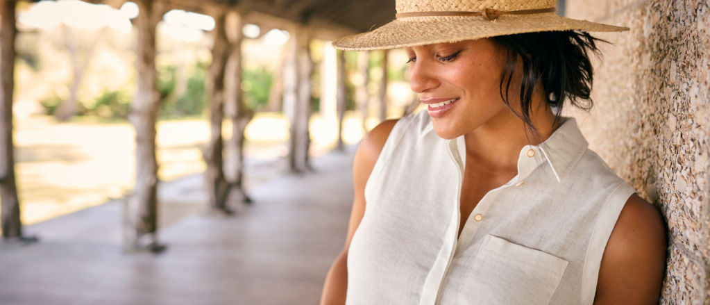 A woman wearing a straw hat and white linen top smiles while leaning against the stone wall of a lodge on a wooden porch.