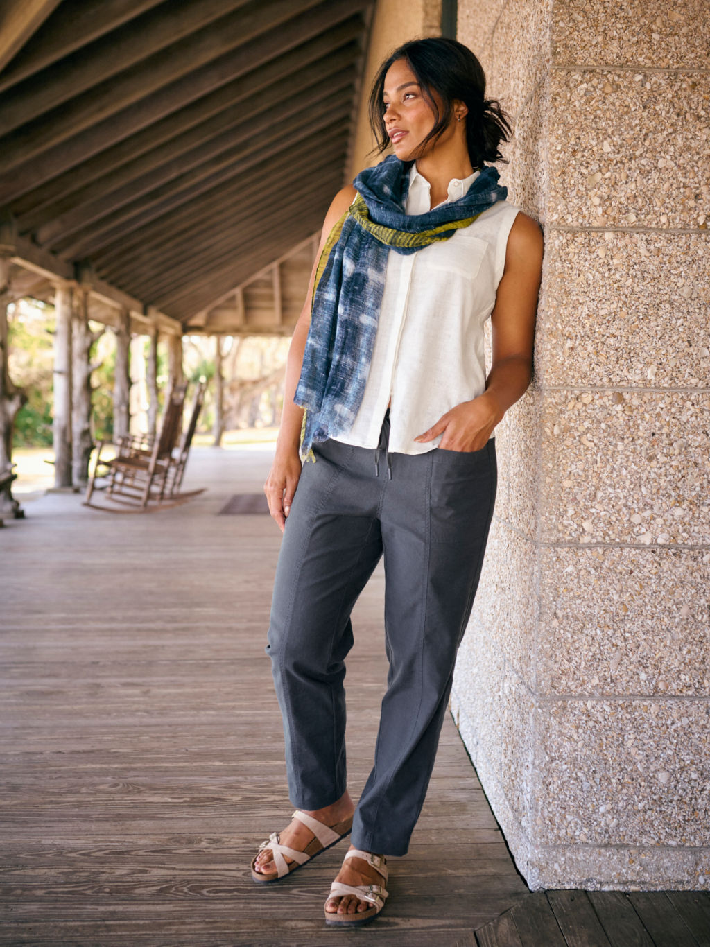 A model wearing a dark blue linen pants, a white linen sleeveless top, and a printed linen scarf leans against a stone wall on a porch.