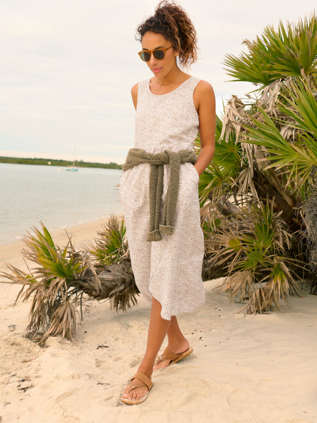 A model wearing a long linen dress, sunglasses, and sandals walks a sandy beach.