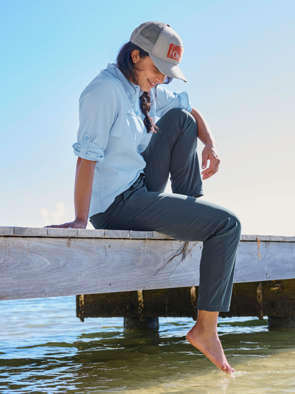 A woman sitting on the edge of a wooden dock with her toes dangling in the water.  Wearing a trucker hat, light blue button down shirt and dark blue pants.