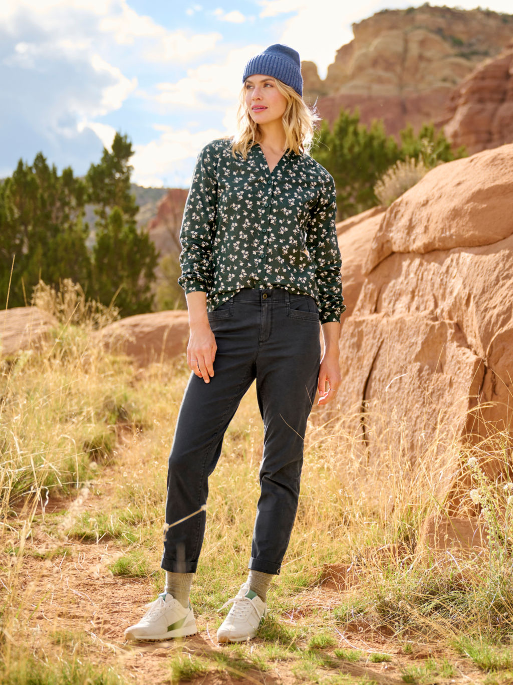 A woman in a green flowered shirt pauses mid-hike in tall dry grass.