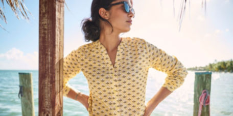 A woman in a printed button-down stands on a dock with the ocean behind her.