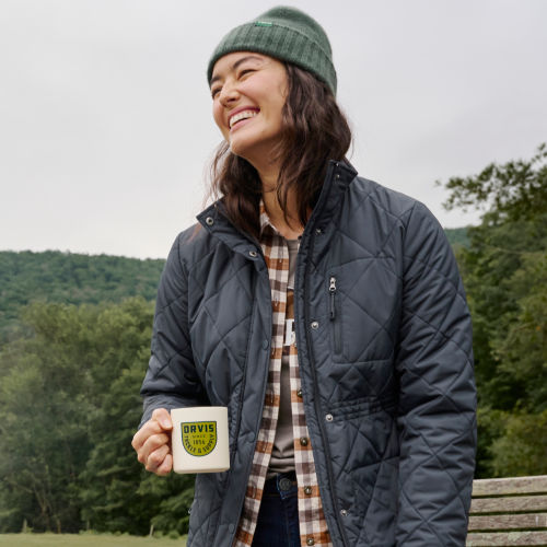 A woman smiles while holding a mug out of doors.