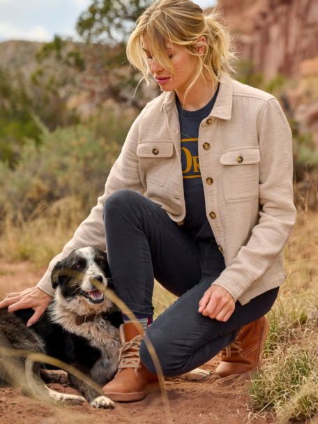 Woman in Here or There Shirt Jacket and Stretch Corduroy Natural Fit Skinny-Leg Pants stops on a desert trail to pet her dog.