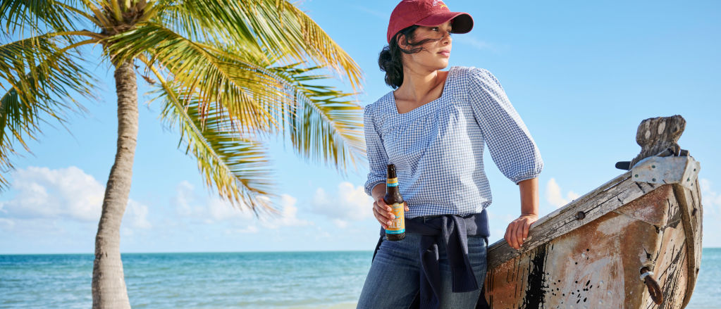 A woman wearing a gingham puff-sleeved blouse and a red baseball cap, holding a beer and leaning on a boat resting on a tropical beach.