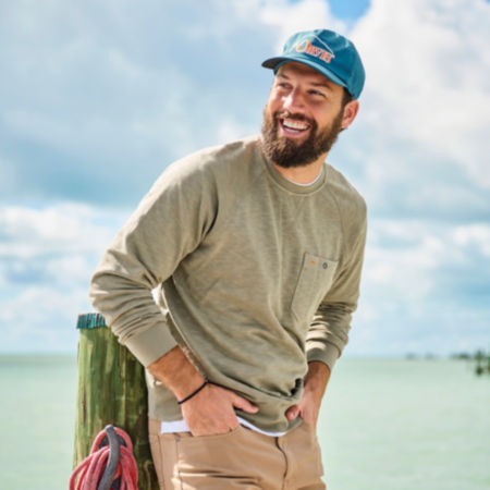 A smiling man in a long-sleeve green tee stands in front of bright tropical ocean.