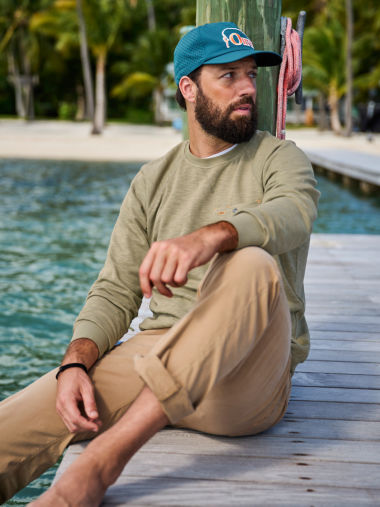 A man sits on a wooden dock wearing a Montana Morning Sweatshirt and Orvis ball cap.