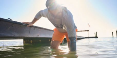 An angler wades knee-deep in green water with one hand on a wooden dock.