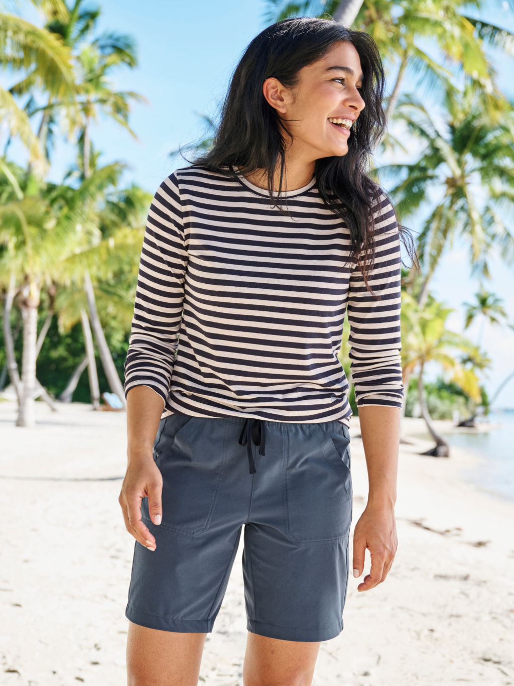 A woman with dark hair wearing a navy and white striped t-shirt and lightweight navy colored shorts standing on a beach.