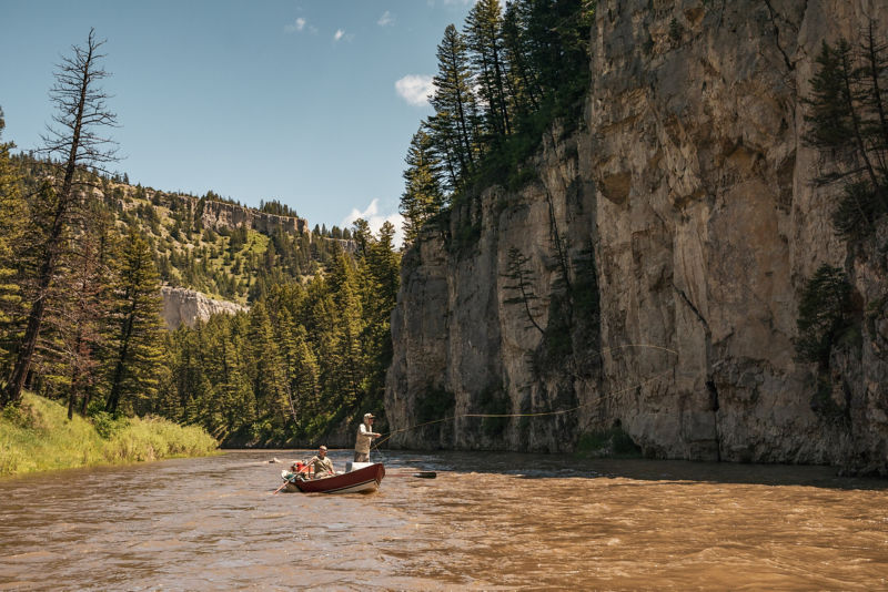 Two people fly fishing in a boat with green trees clinging to sides of rocky cliffs on either side.