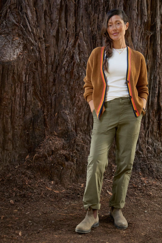Woman standing in front of a tree wearing brown sweater, white t-shirt, and green pants.
