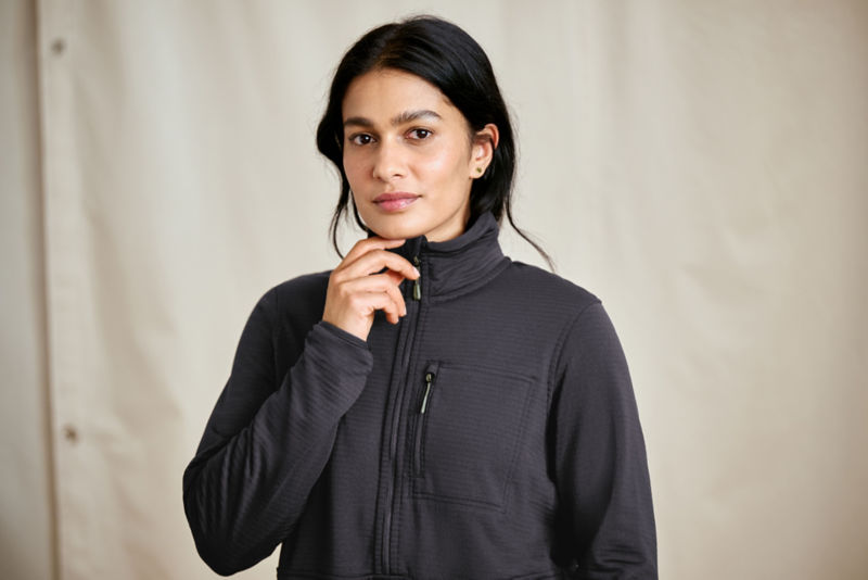 A model stands in front of a canvas drop cloth wearing a black softshell sweatshirt zipped up to her chin.