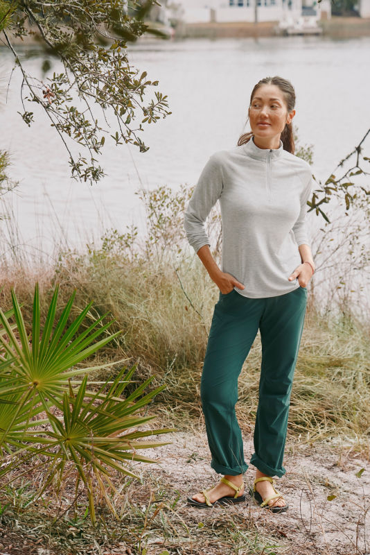 Woman wearing grey quarter-zip and green pants standing next to palm trees.