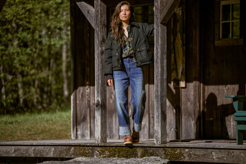 Model wearing olive barbour jacket with silk shirt and jeans standing on front porch