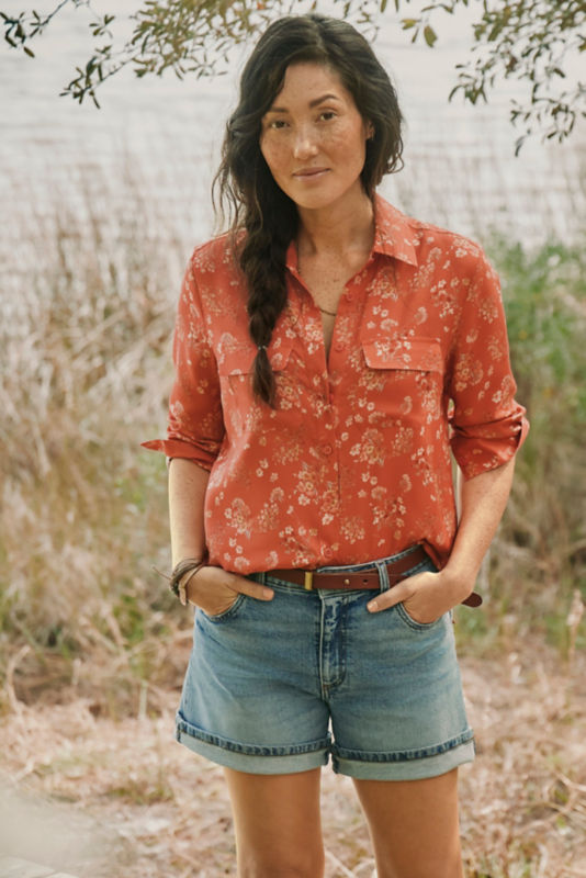Woman wearing red shirt and jean shorts standing in front of a body of water.