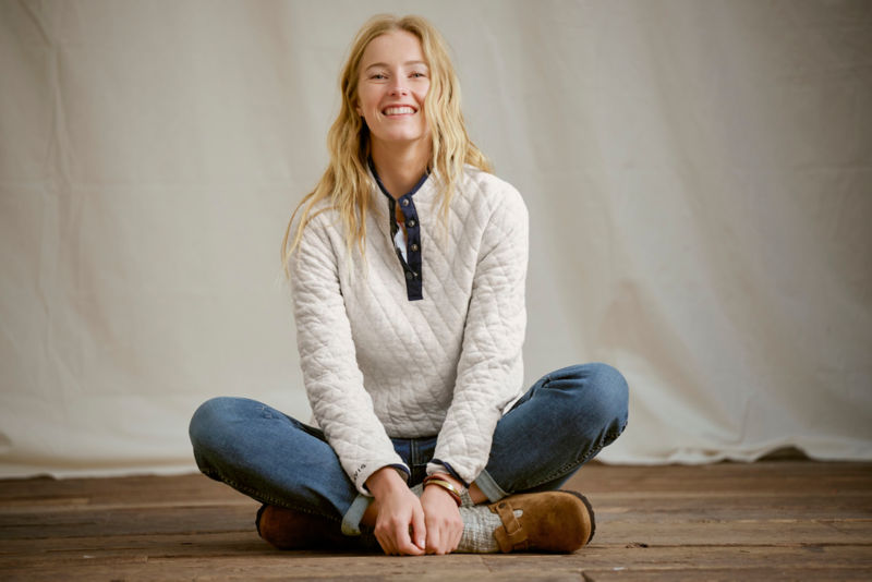 Woman wearing grey quilted pullover sweatshirt and jeans in a studio.