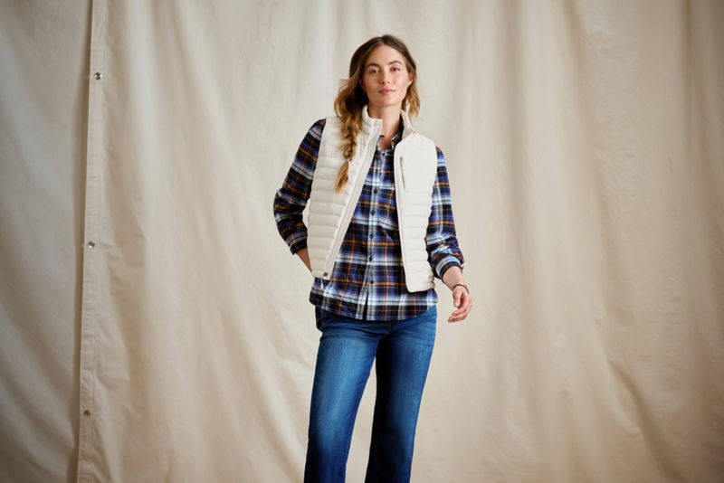 A woman wearing a cream puffy vest over a blue plaid shirt with blue jeans.
