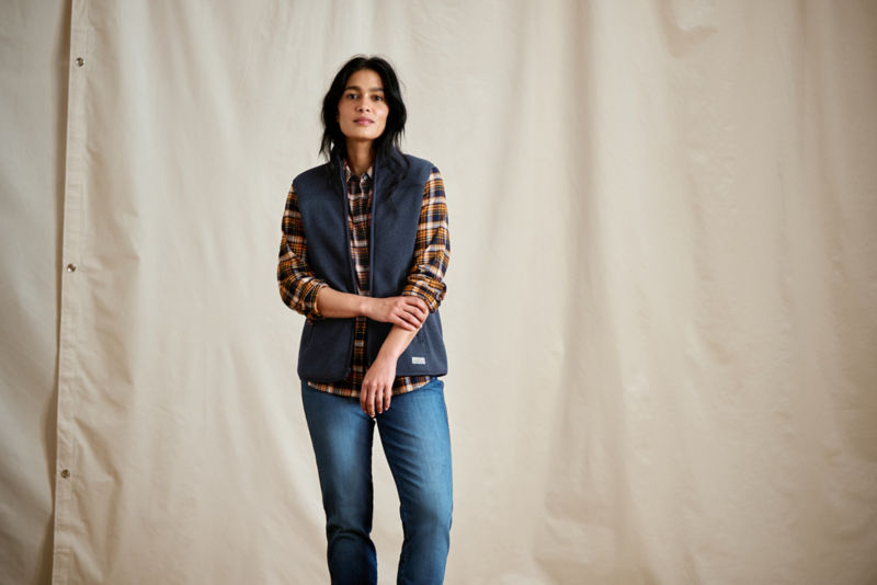 A woman in a plaid flannel shirt with a navy vest layered on top, pulling her sleeve up.