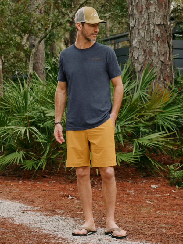 A model wearing a navy tee and gold shorts stands on a gravel path.